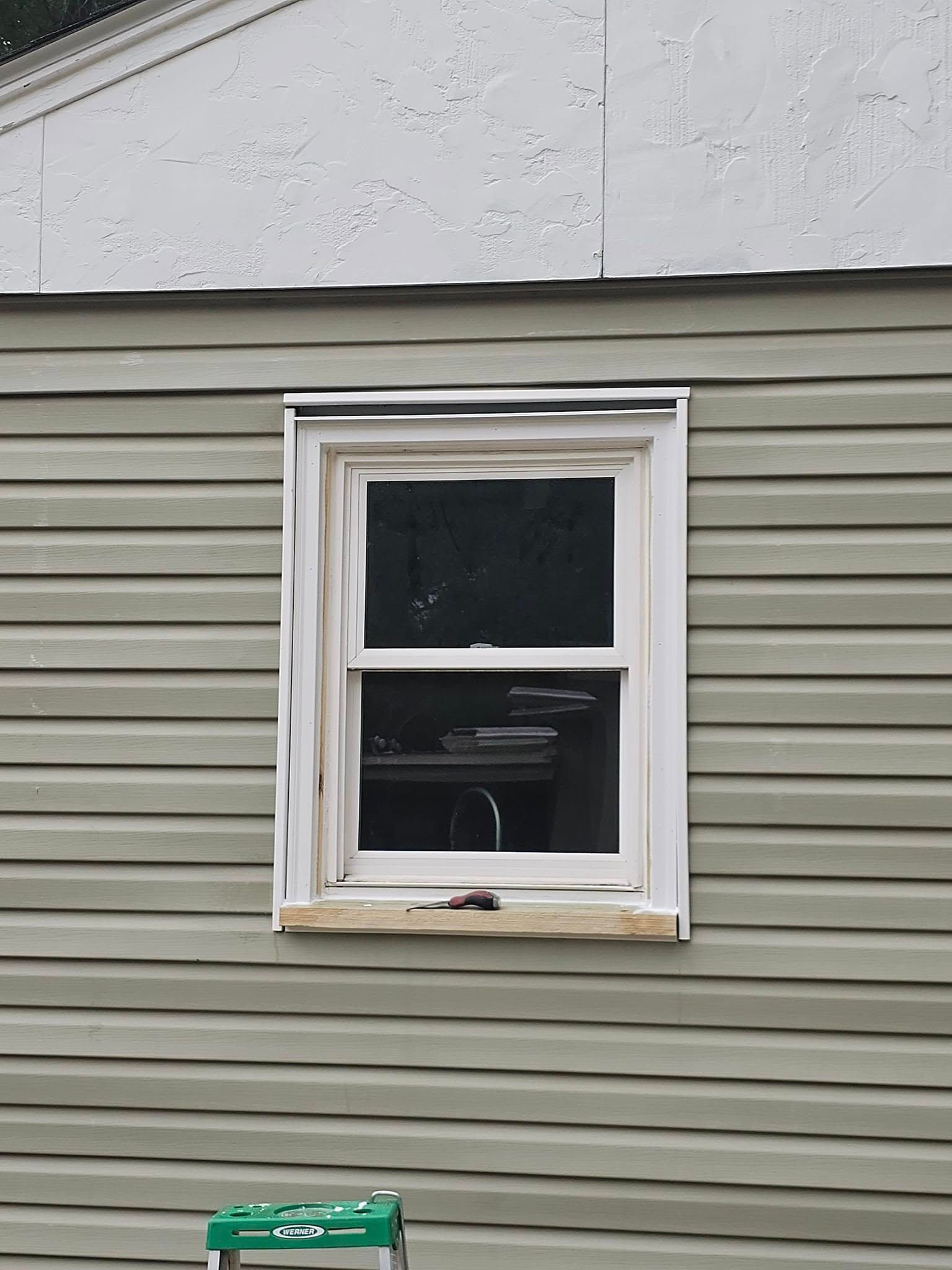 Window in a house with green siding, surrounded by white trim. Green ladder visible in front.