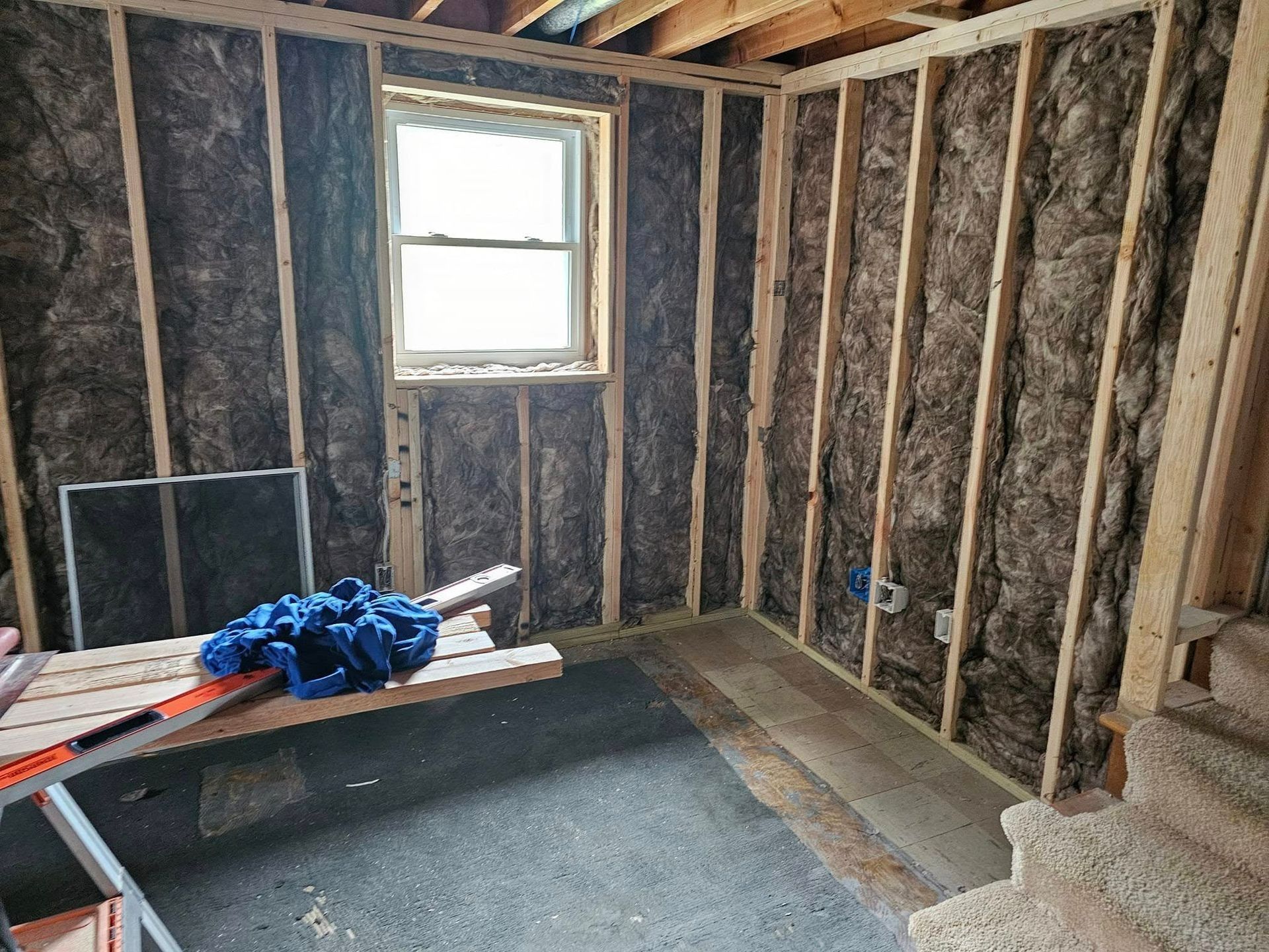 Interior of a room under construction, with wood frame walls and insulation, window visible, construction materials present.
