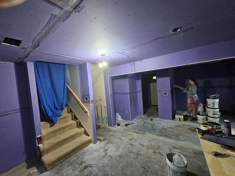 Room being painted purple, with woman painting the wall. Staircase with blue curtain, and paint buckets are visible.