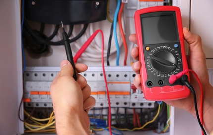 A person is holding a multimeter in front of an electrical box.