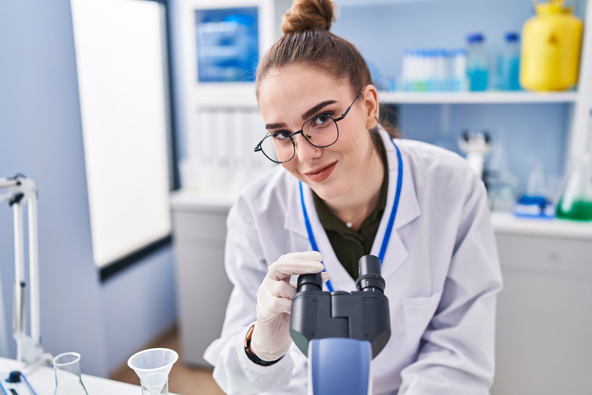 Mujer con bata de laboratorio utilizando un microscopio, sonriendo en un entorno de laboratorio.