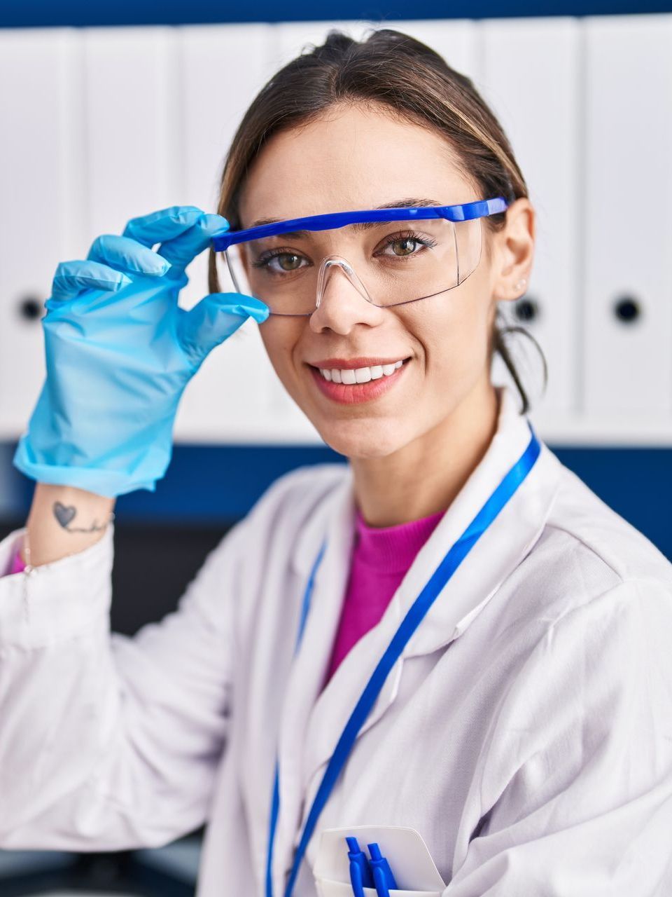 Mujer con bata de laboratorio y guantes ajustándose gafas de seguridad, sonriendo en un entorno de laboratorio.