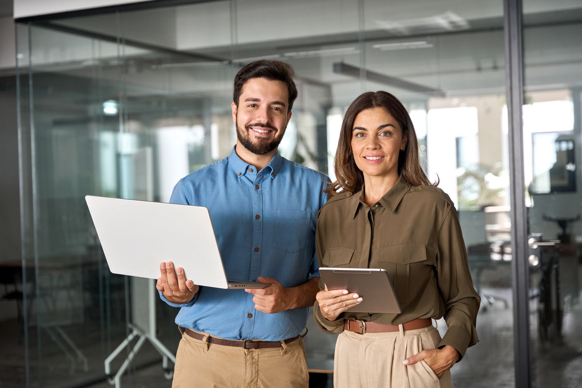 A man and a woman are standing next to each other in an office holding a laptop and a tablet.