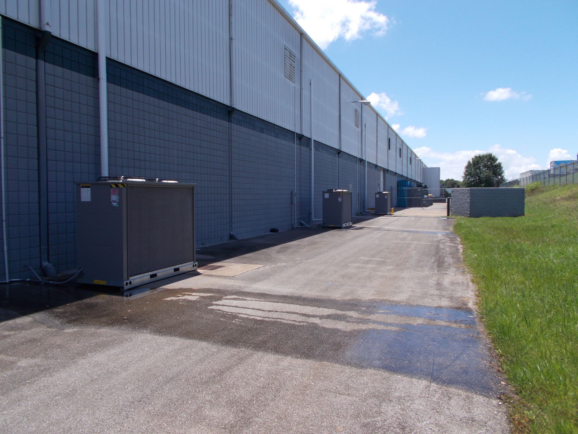Long gray warehouse wall beside a paved service road and grassy field under a blue sky