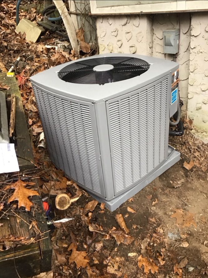 Outdoor air conditioning unit beside a house, surrounded by fallen leaves and dirt.