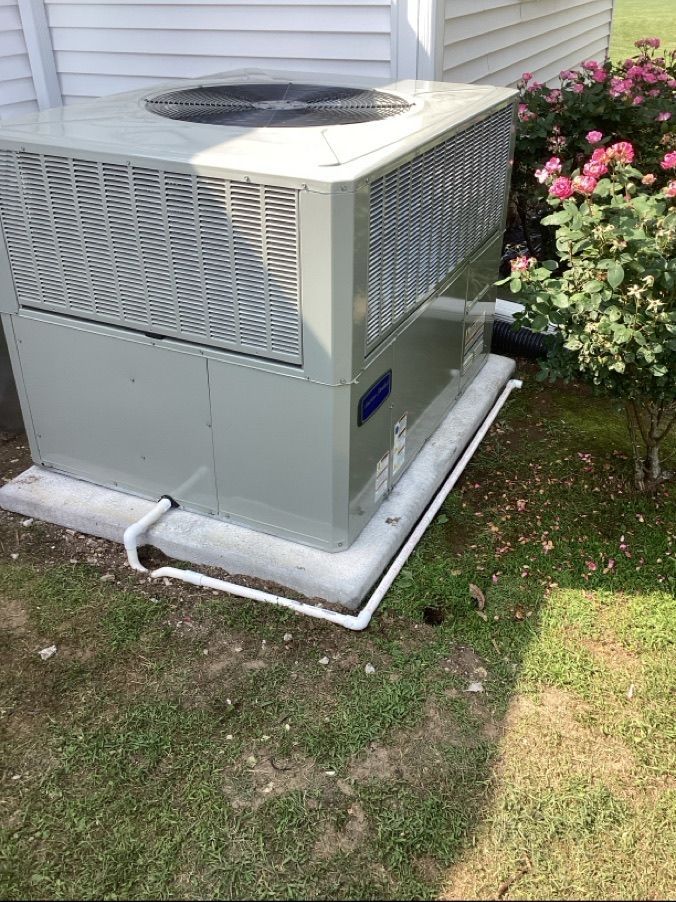 Outdoor air conditioner unit beside a house, with white piping and flowers nearby