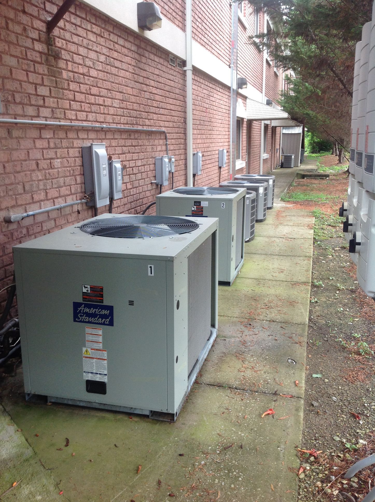 Row of outdoor HVAC units along a brick alley beside a building, on a damp concrete path.