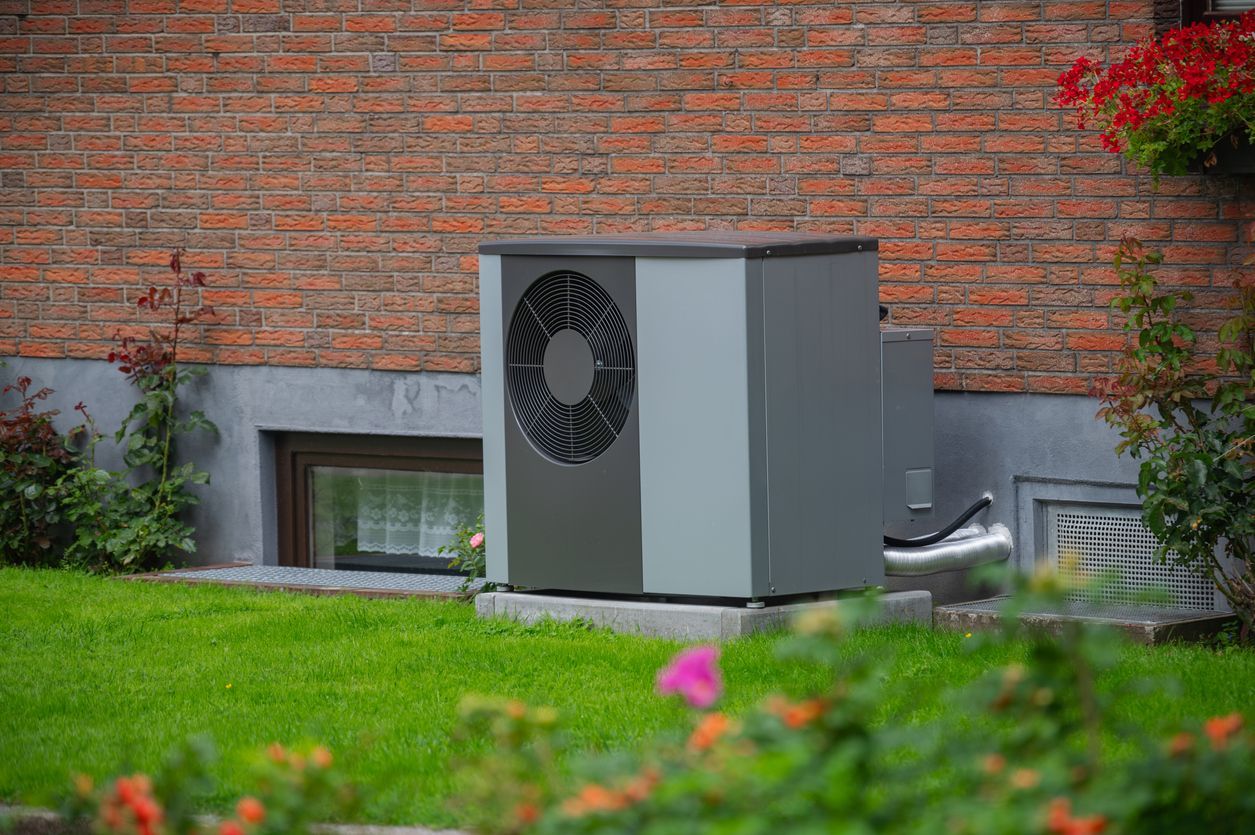 Outdoor HVAC unit beside a brick building, framed by green grass and flowers