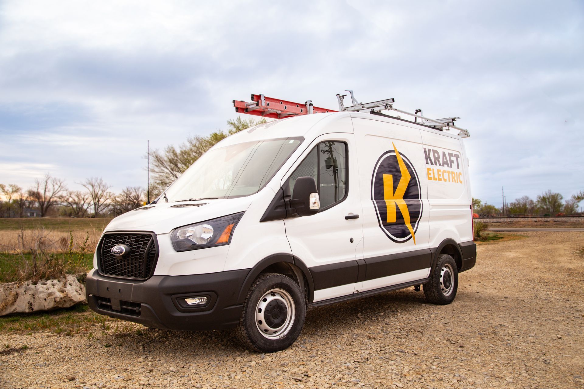 White service van with ladder racks and logo parked on a gravel lot under cloudy sky