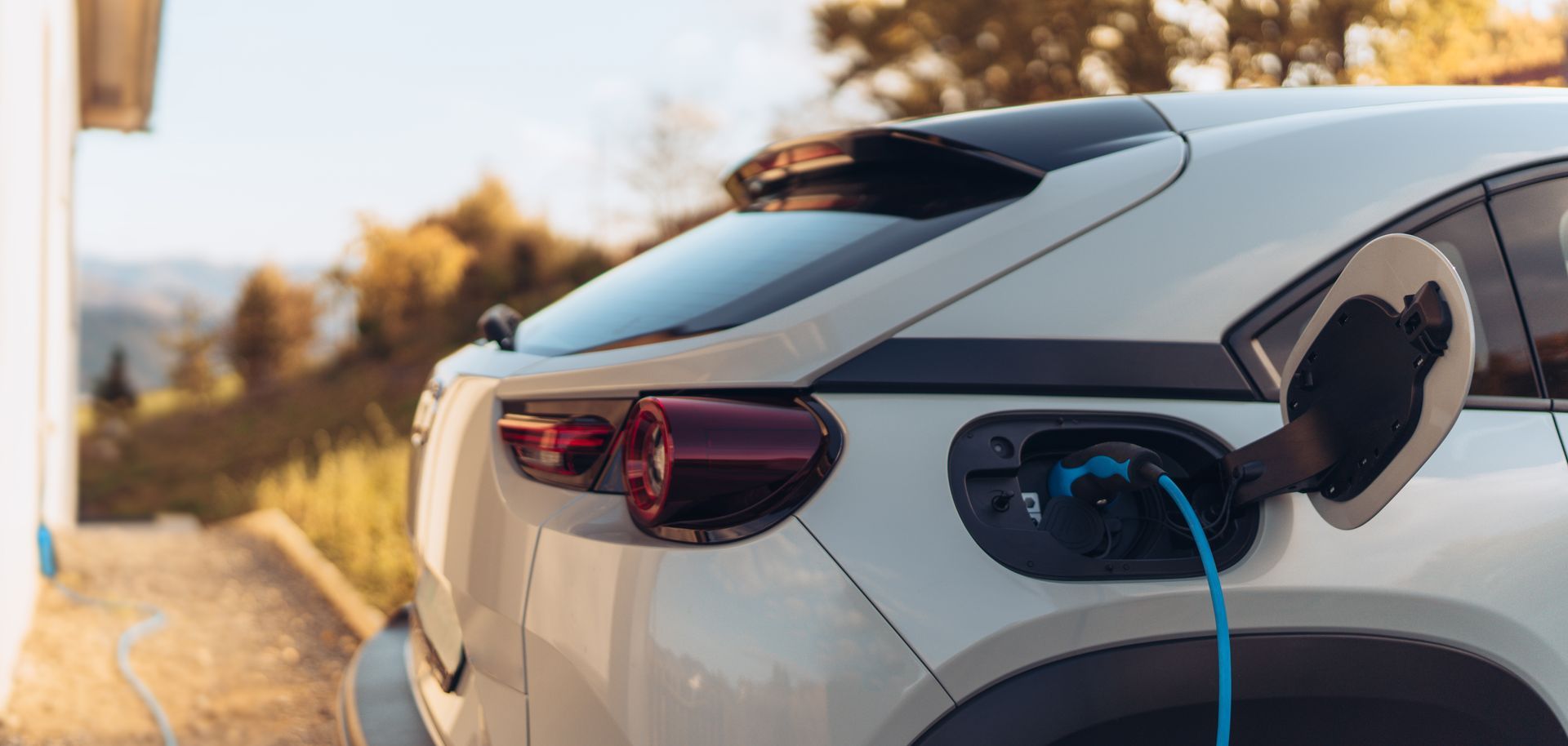 White electric car charging outdoors at sunset, with a blue cable connected to the rear port