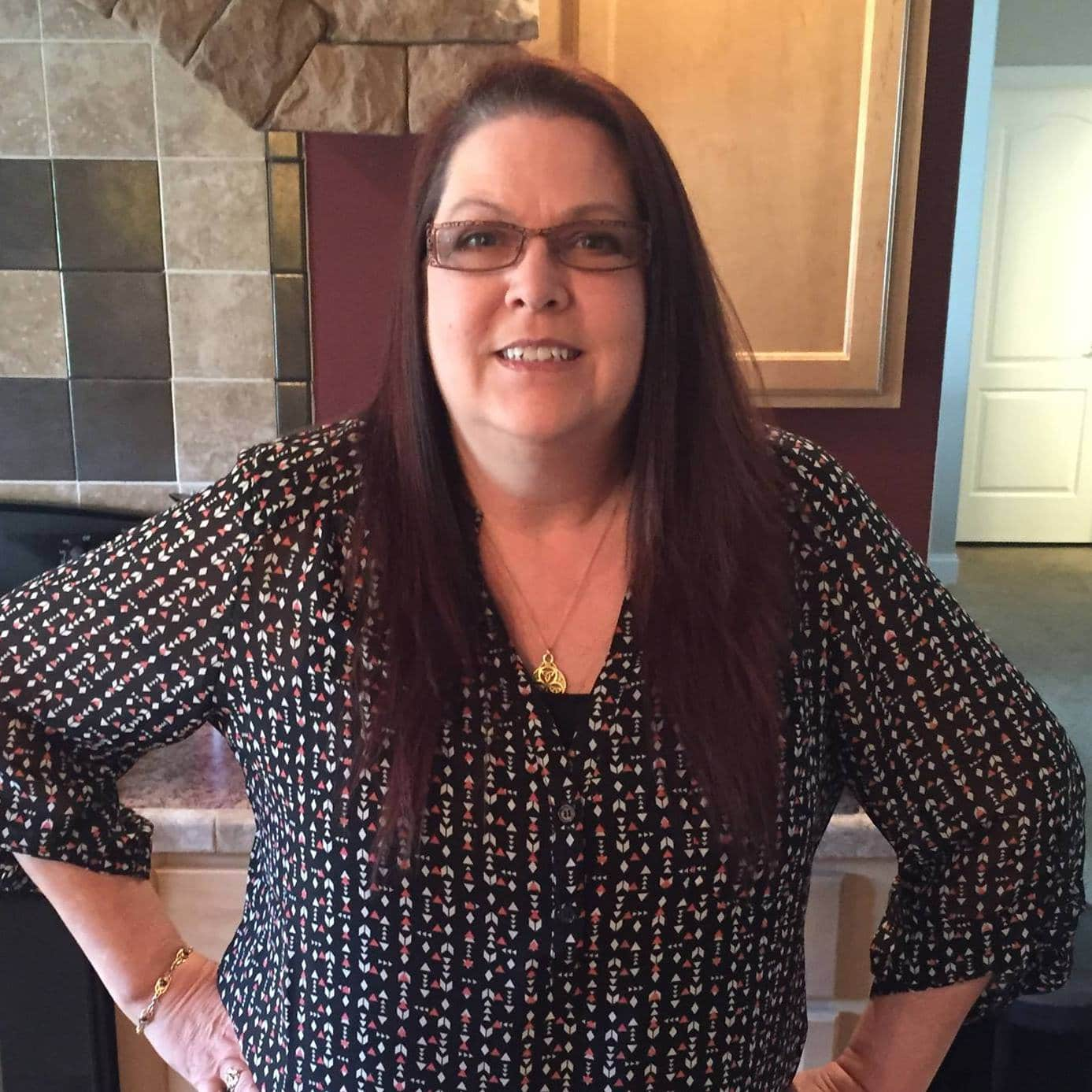 Woman with glasses, hands on hips, wearing a patterned top, in front of a fireplace and a kitchen counter.