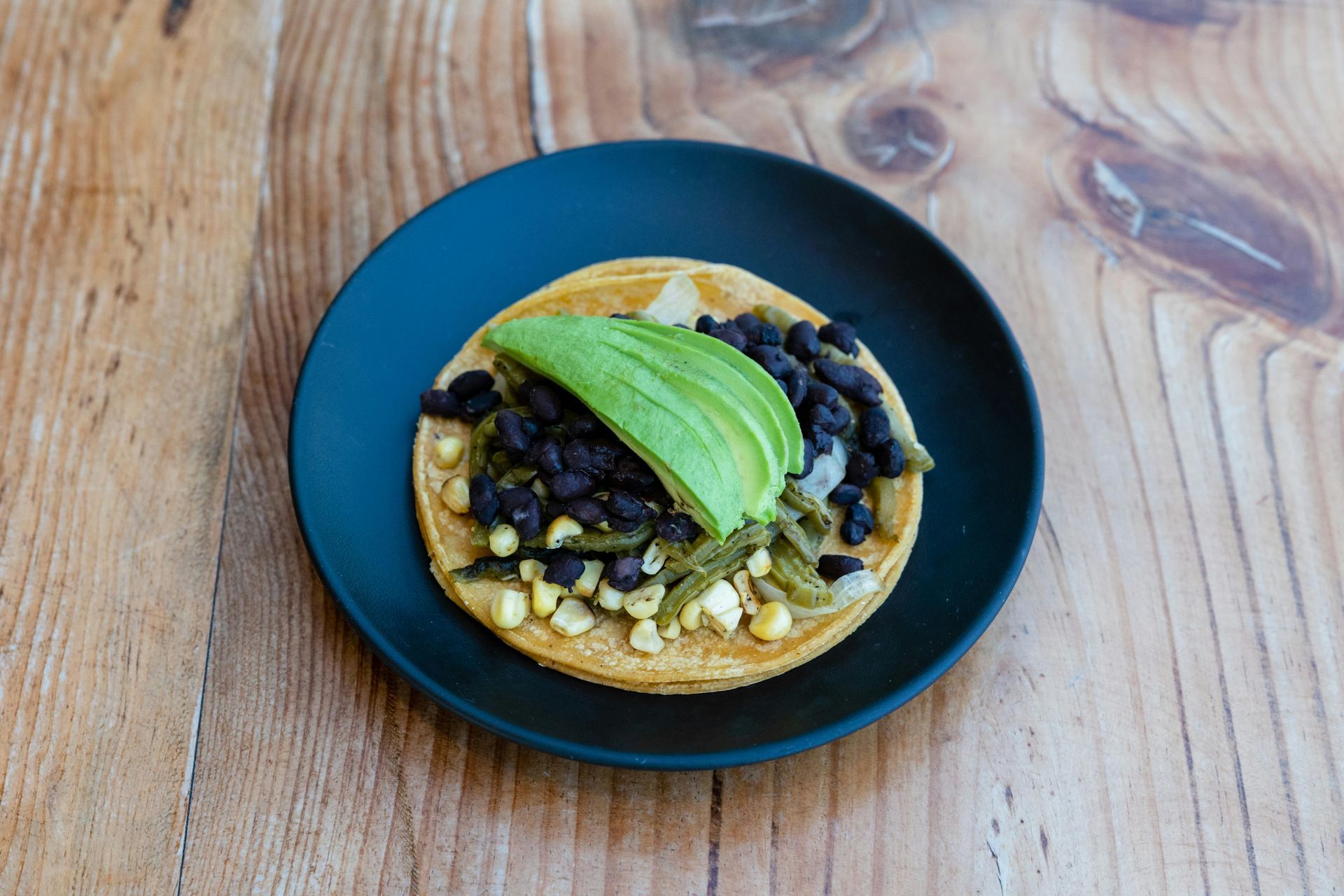 Taco on black plate: corn tortilla, avocado, black beans, corn, and green chiles on wood table.