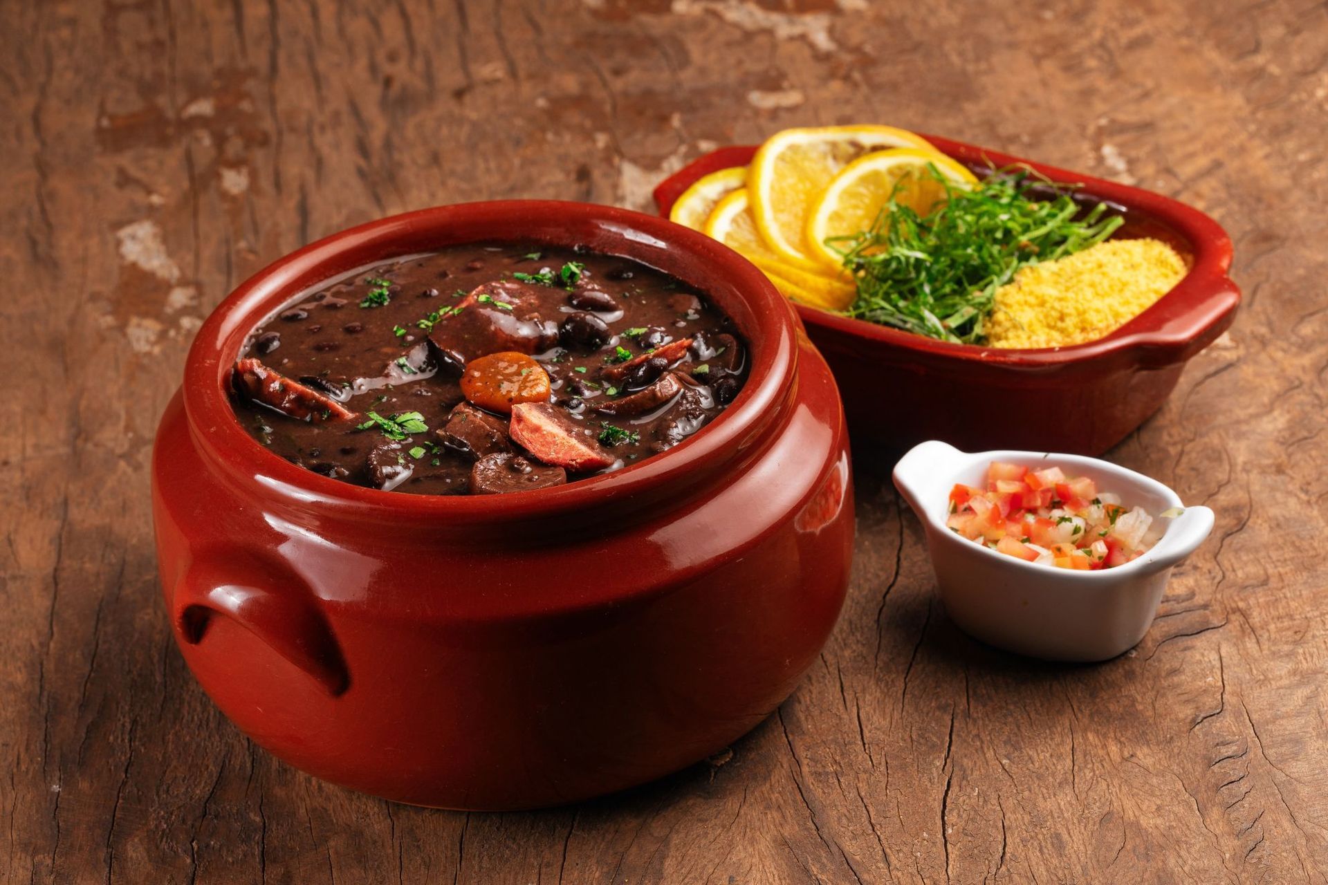 Feijoada stew in a ceramic pot with sides of orange slices, farofa, and salsa on wooden table.