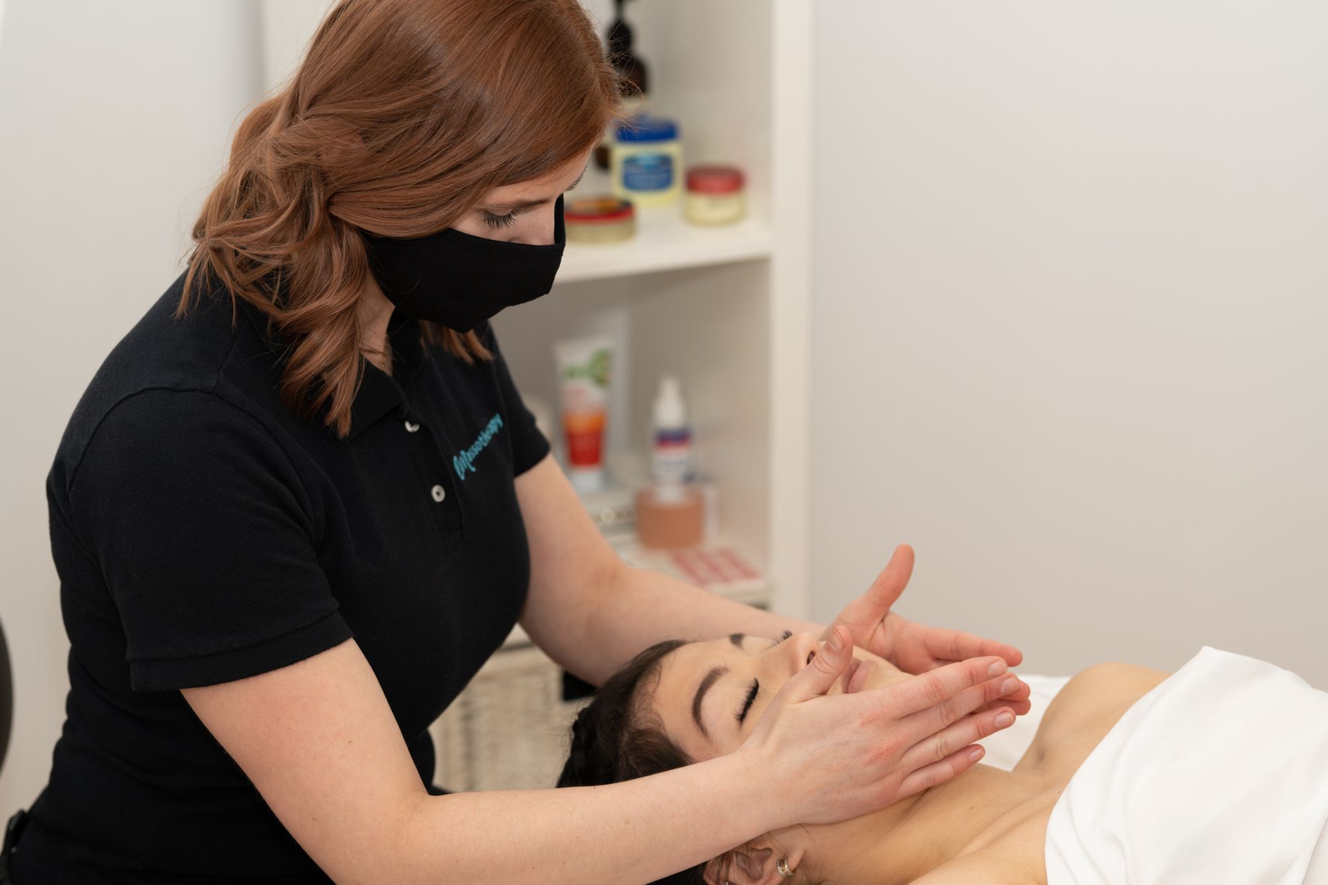 Une femme masquée donne un massage du visage à une personne allongée dans un cadre de type spa.