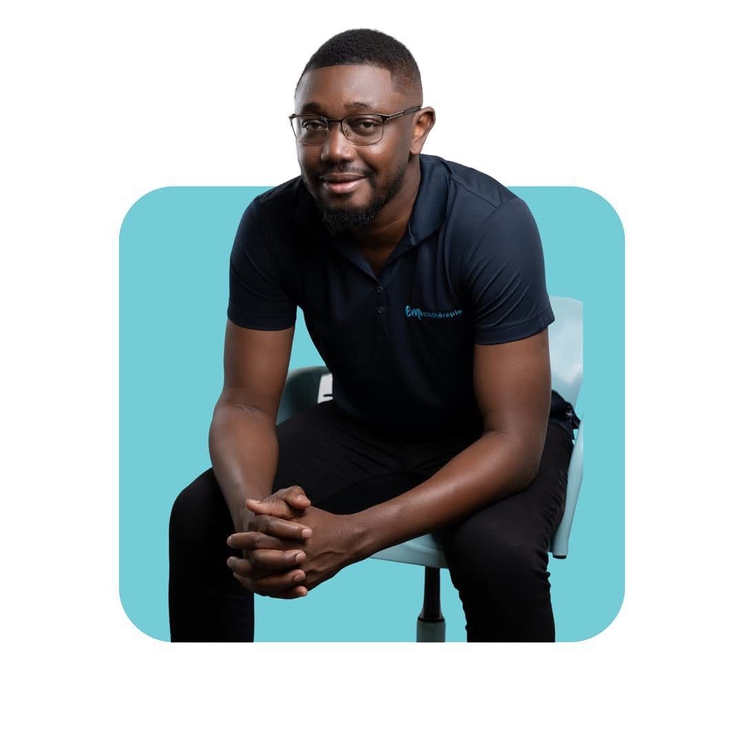 Homme en chemise et pantalon noirs, assis sur une chaise, souriant, les mains jointes. Fond bleu.