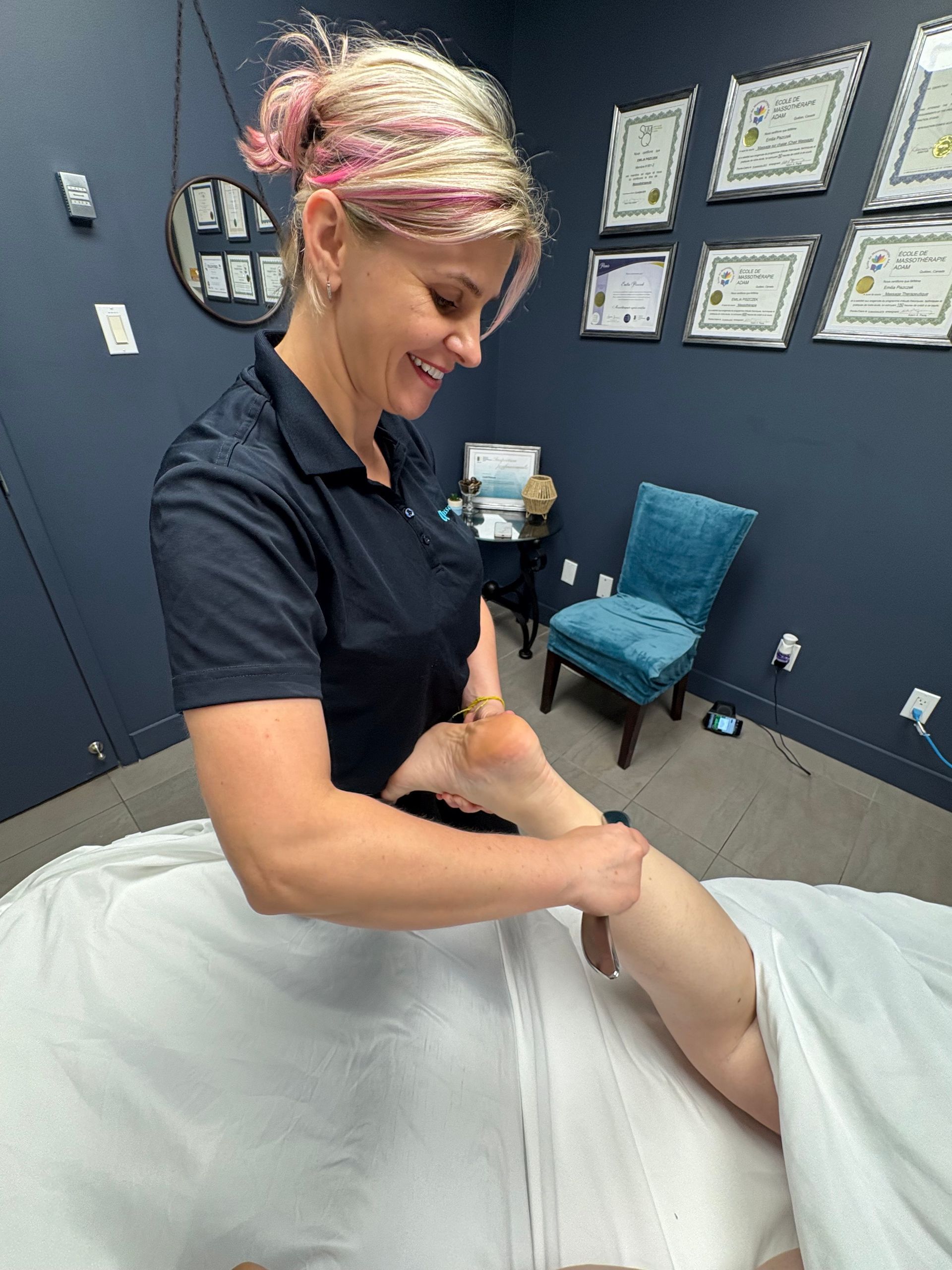 Une femme masse les jambes dans une salle de massage. Elle sourit. Des murs bleus et des certificats sont visibles.