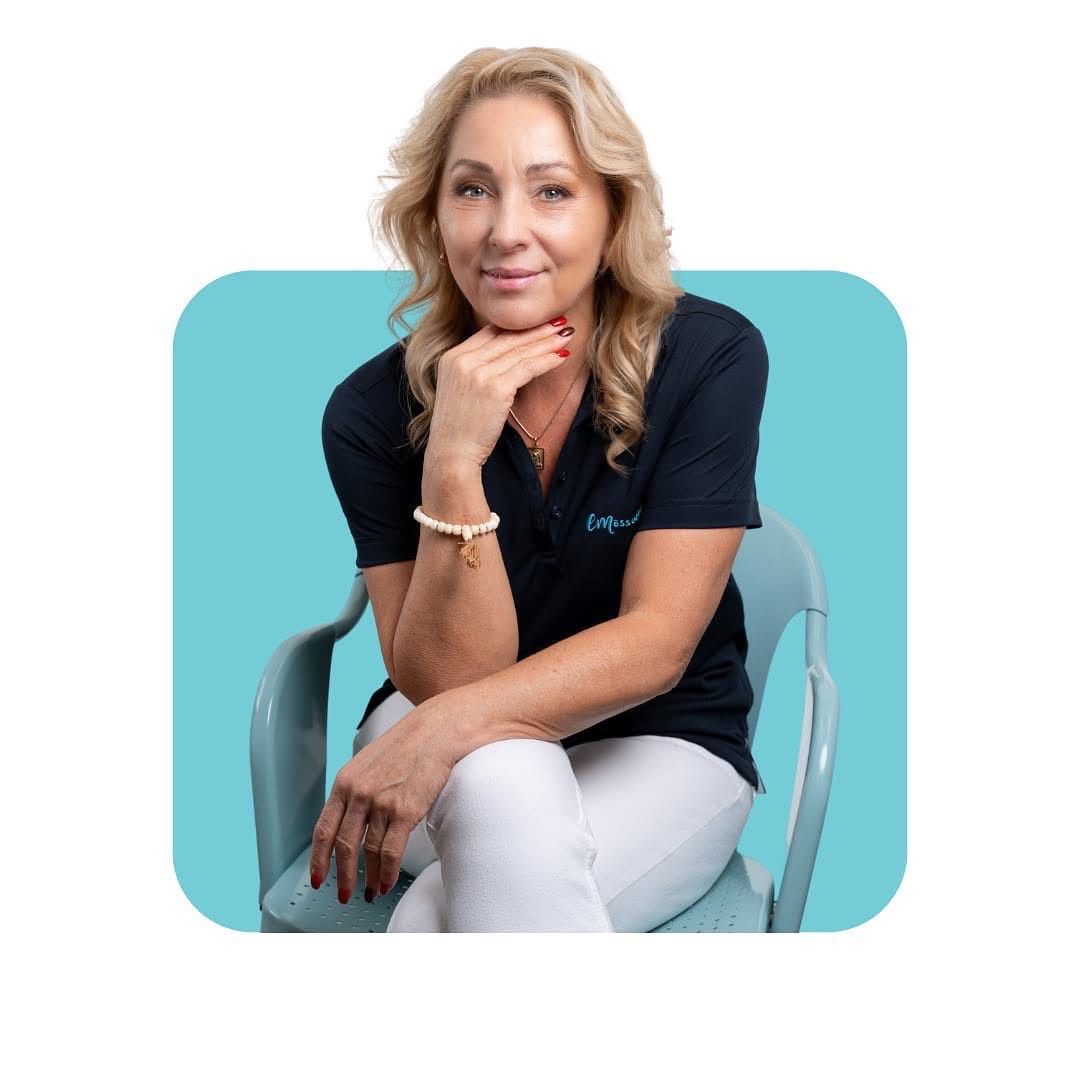 Femme en chemise bleu marine et pantalon blanc, assise sur une chaise bleu sarcelle, la main sur le menton. Fond bleu.