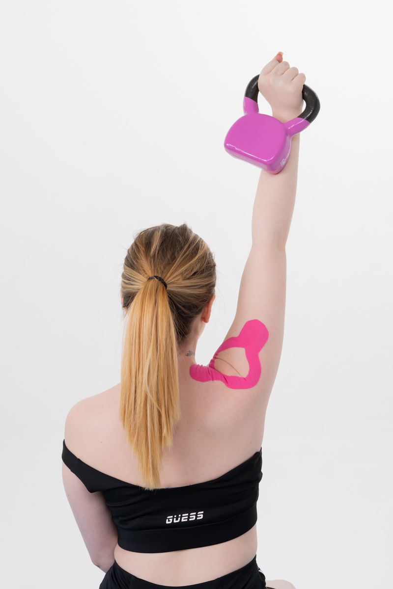 Femme tenant une kettlebell rose au-dessus de sa tête ; ruban de kinésiologie rose sur le bras ; portant un haut noir.