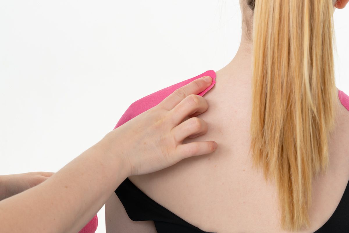 Personne avec du ruban de kinésiologie rose sur l'épaule, la main d'une autre personne touchant le ruban, fond blanc.