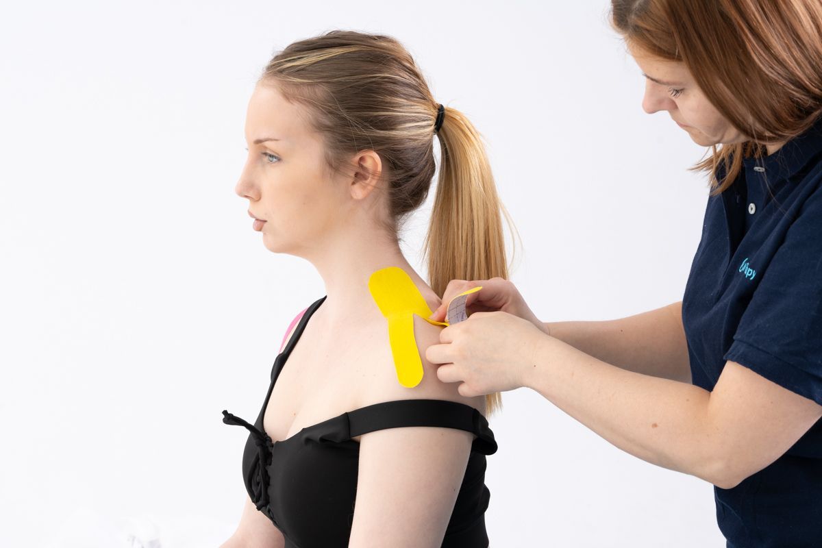 Une femme se fait appliquer du ruban de kinésiologie sur l'épaule par une autre personne.