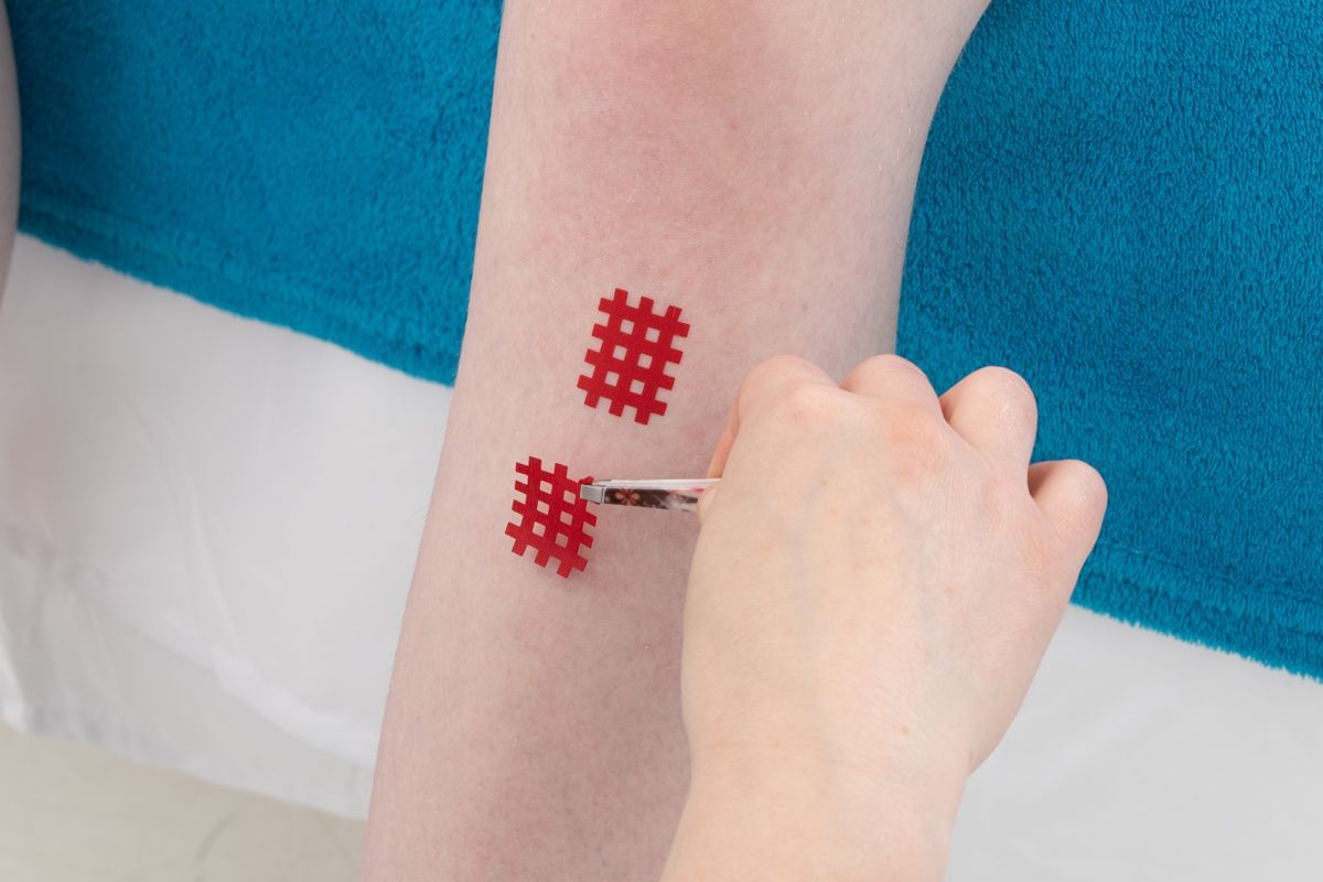 Main appliquant un bandage rouge en forme de treillis sur la jambe, à proximité de deux bandages similaires sur la peau.