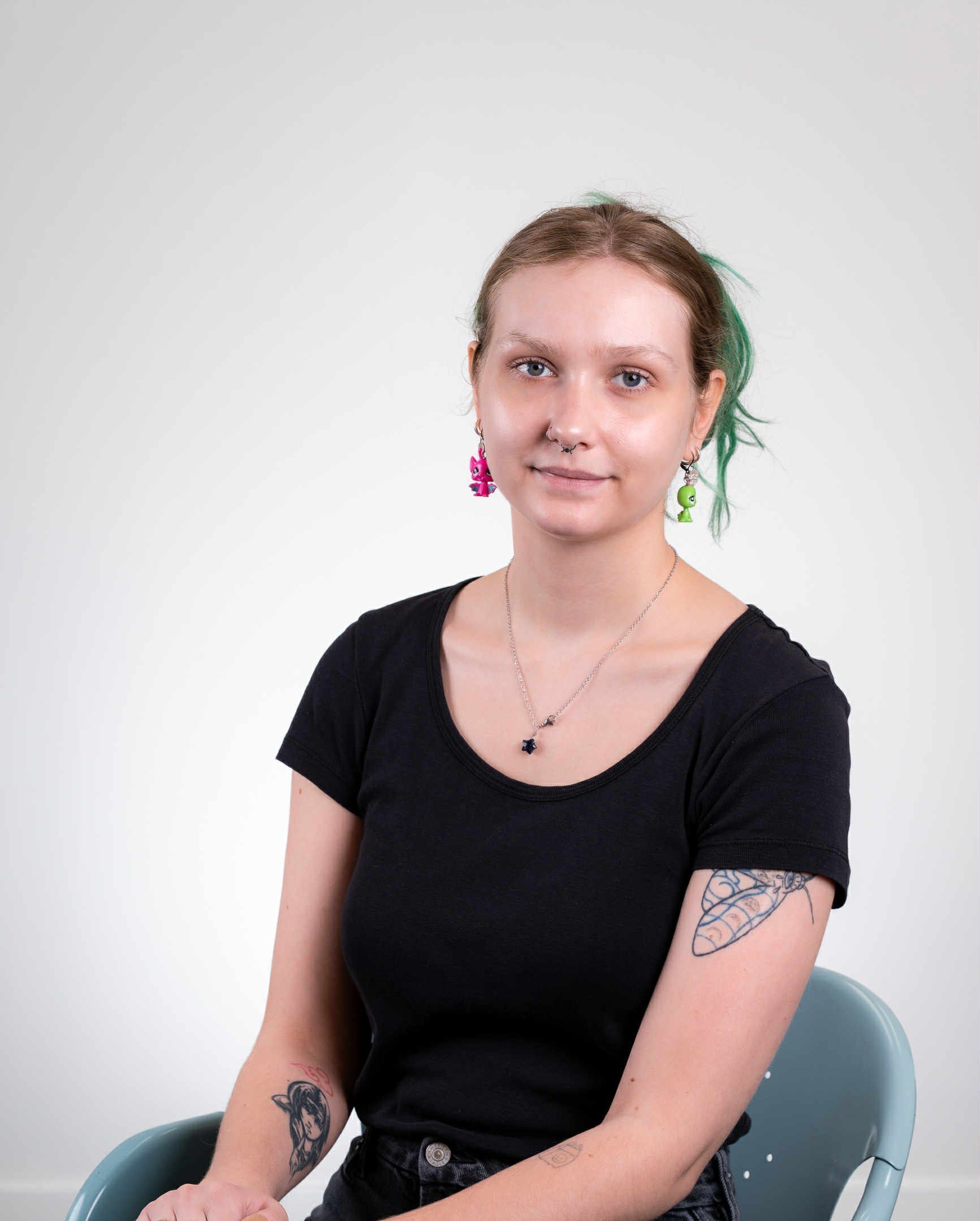 Femme aux cheveux verts, chemise noire et piercing au nez, assise.