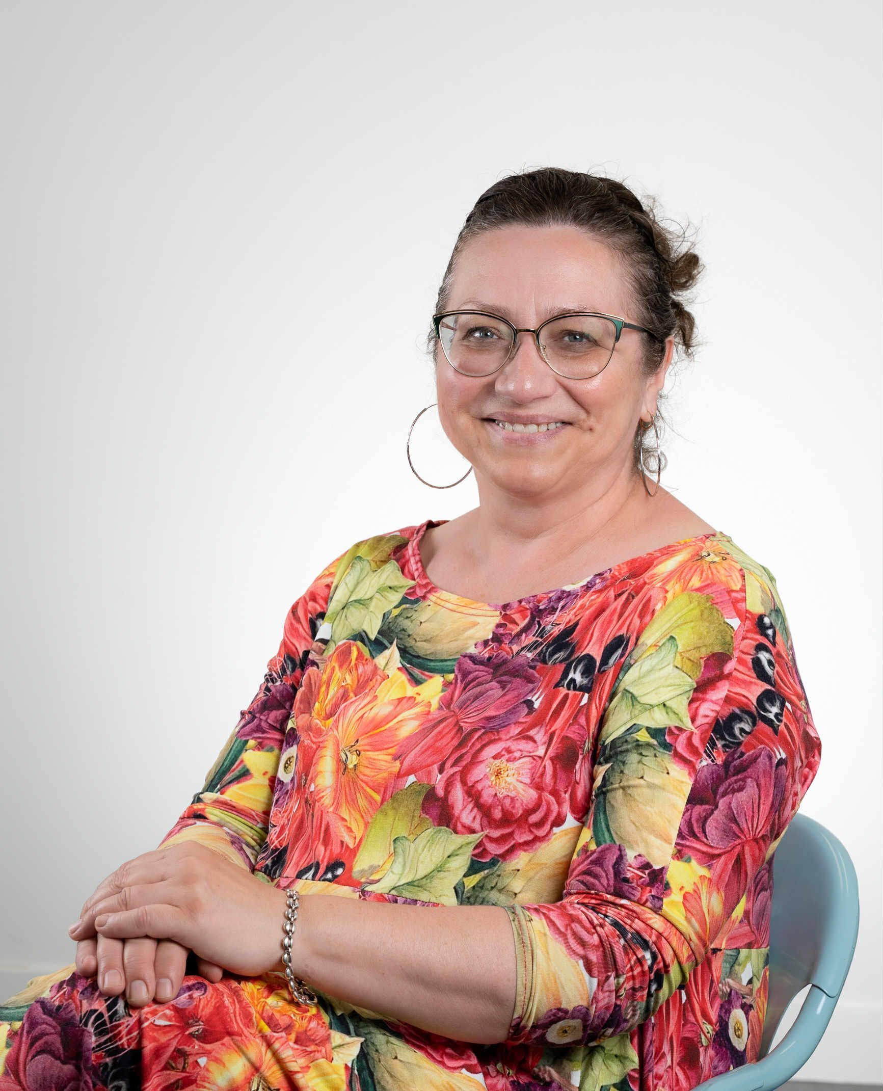 Une femme portant des lunettes et un haut fleuri sourit assise.