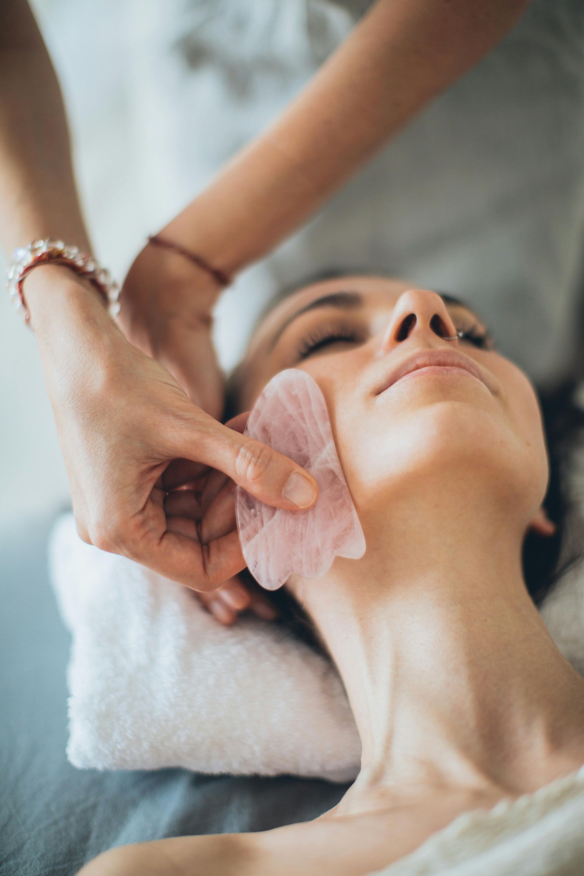 Des mains utilisent un outil Gua Sha rose sur le visage d'une personne allongée sur une serviette blanche.