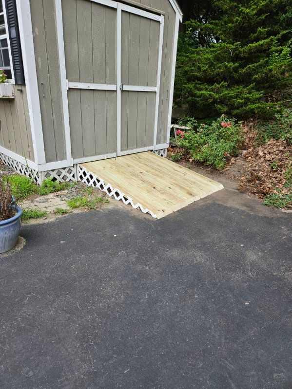 Wooden ramp leads to a shed. Ramp is on asphalt next to a flower bed.