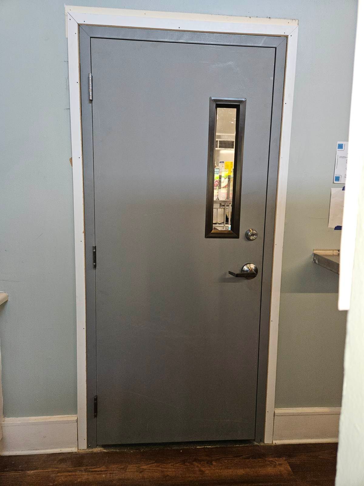 Gray door with small window, black frame, and white trim. Set in a light blue room with wooden floor.