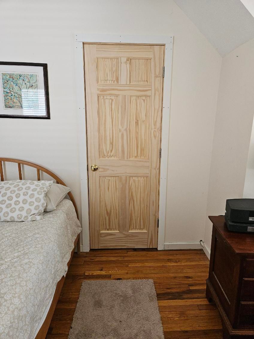 Wooden door set in a white-walled room with a bed and dresser; wood floor.