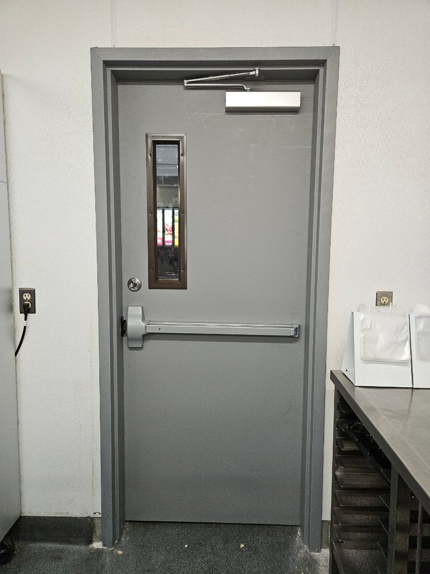 Gray metal emergency exit door with push bar, window, and white wall.