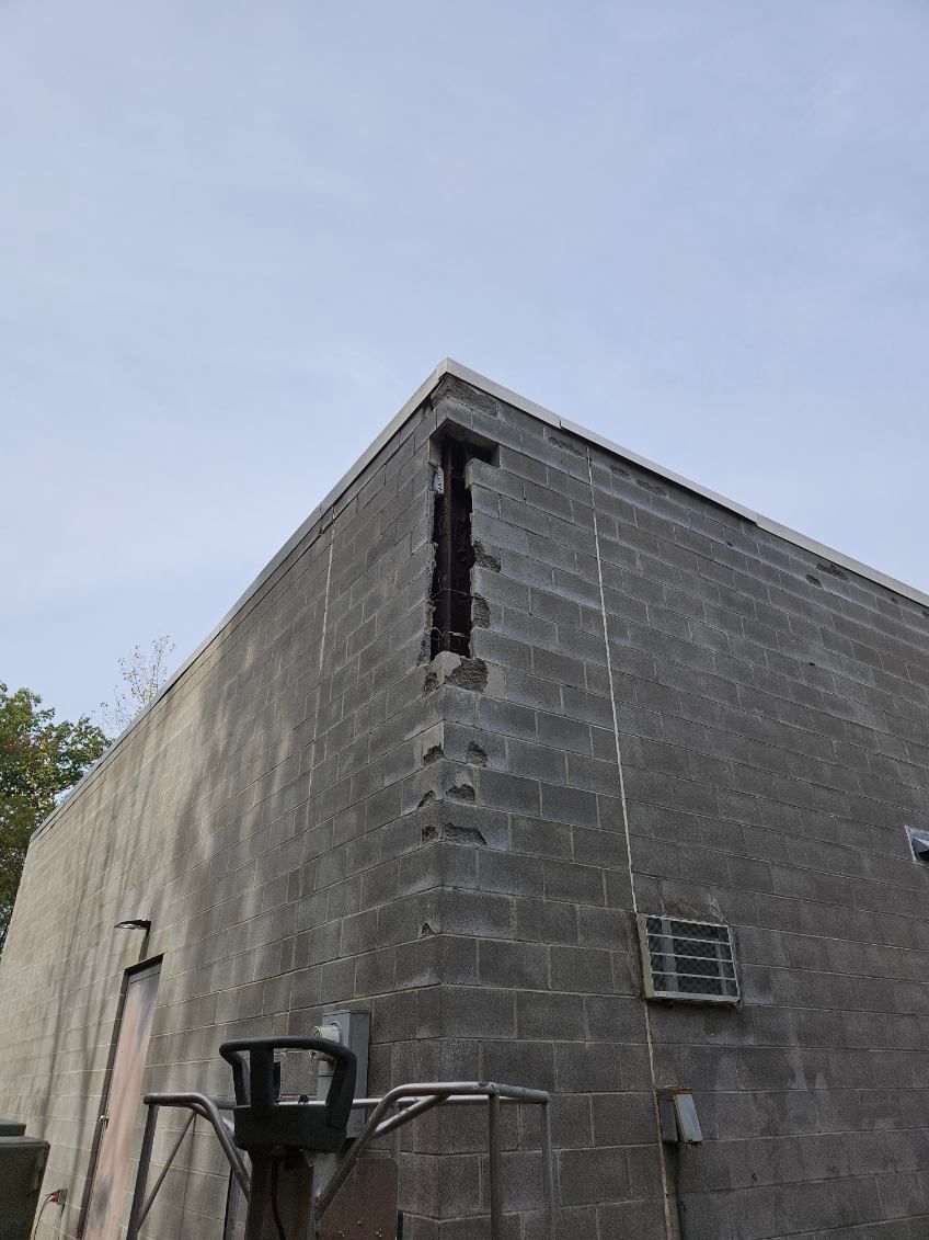 Corner of a gray cinder block building with damage, against a light sky.