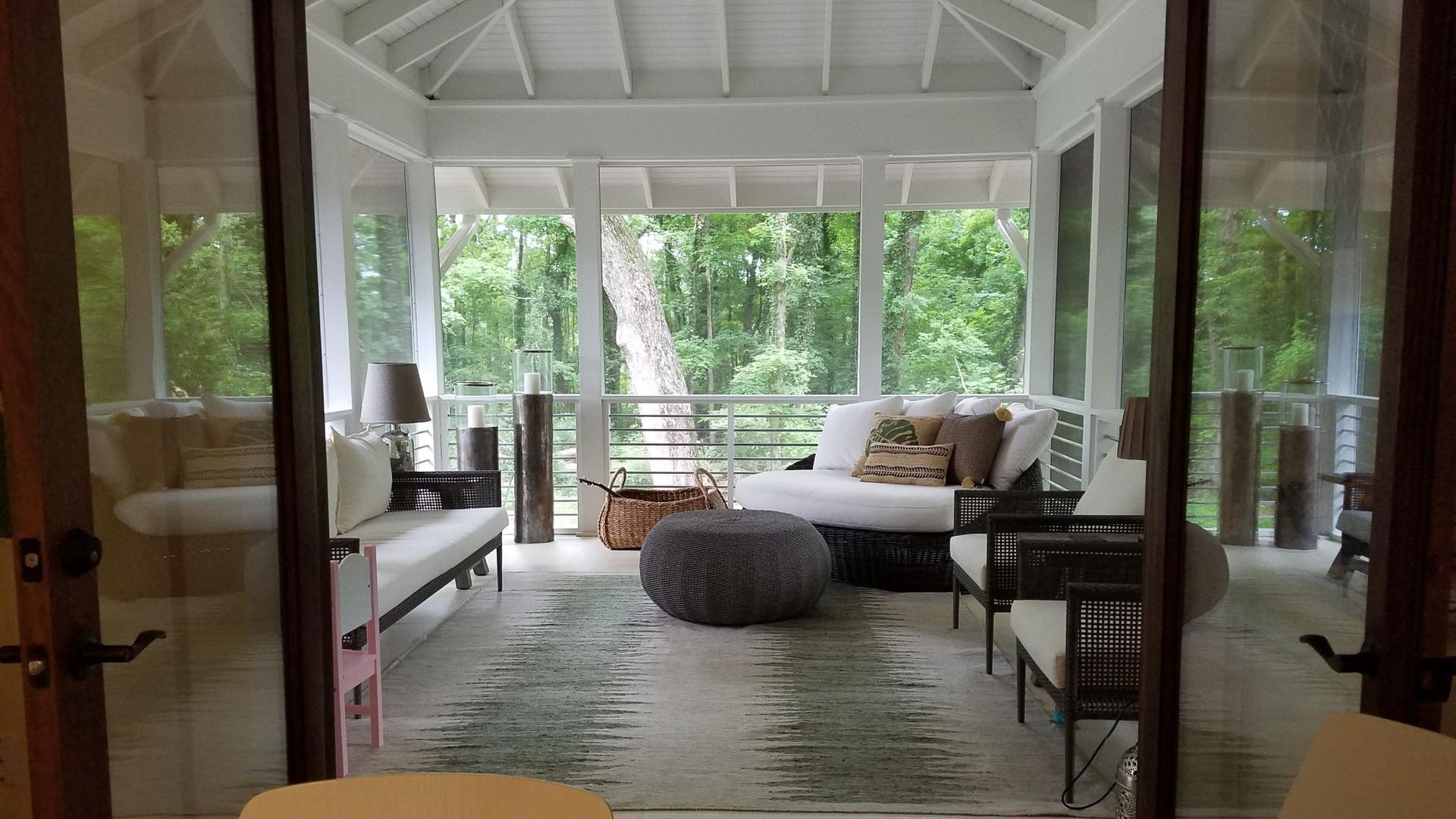 Bright porch with white wicker sofa, ottoman, and dining chairs overlooking green trees.