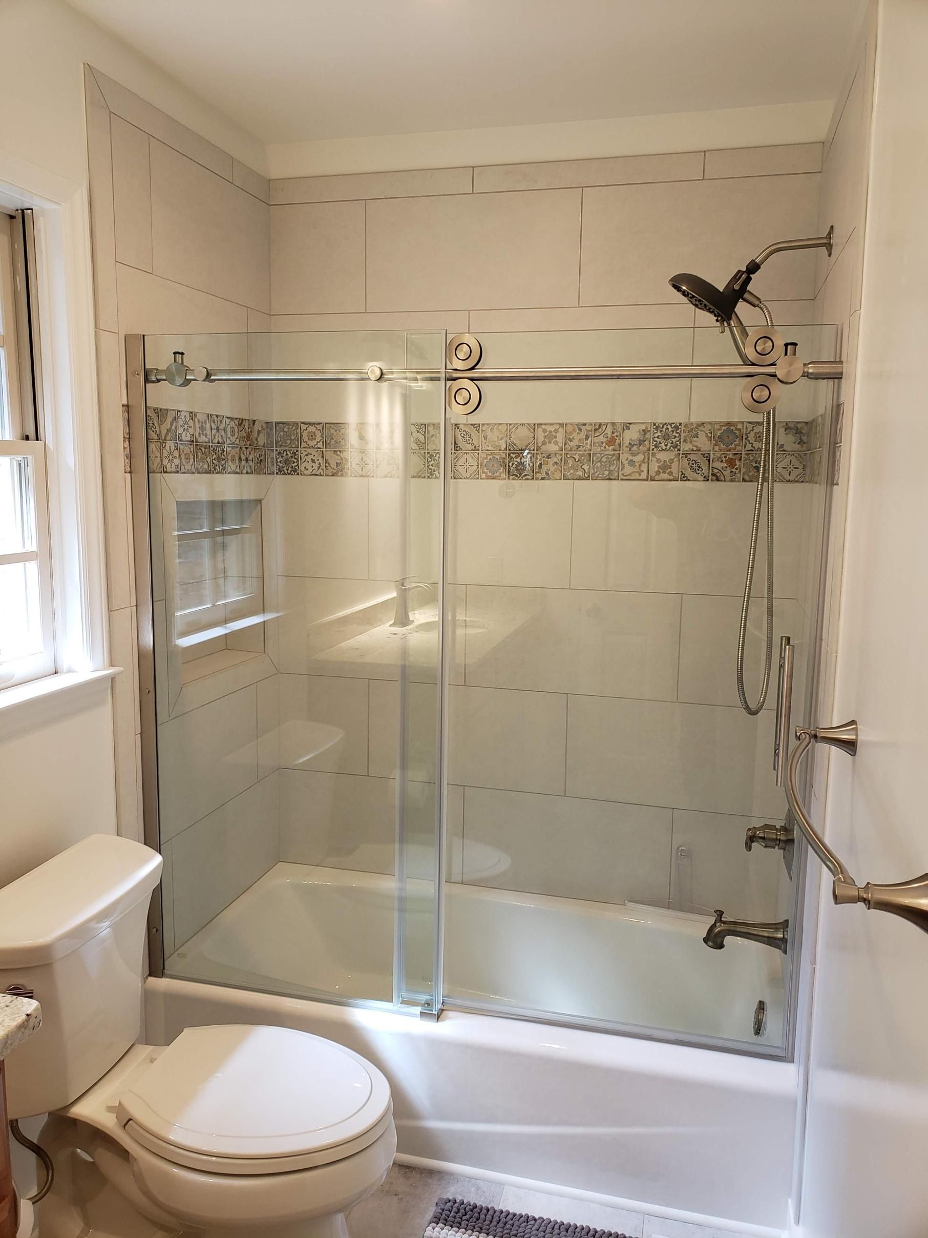 Bathroom with glass shower enclosure, beige tile accents, toilet, and window on the left.
