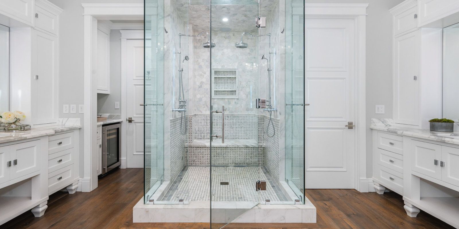Modern bathroom with a glass walk-in shower centered between white double vanities and wood floors