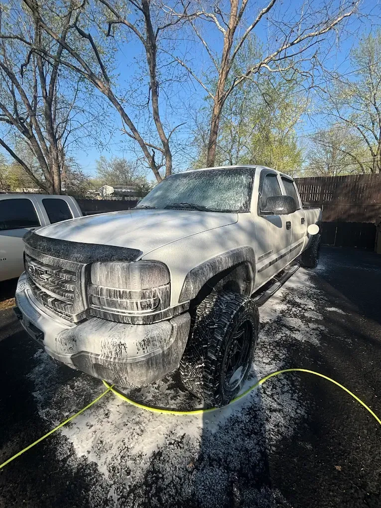 White pickup truck covered in thick foam at a car wash, with trees and parked vehicles in the background
