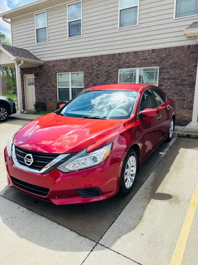 Red Nissan sedan parked in a lot beside a brick and beige apartment building.