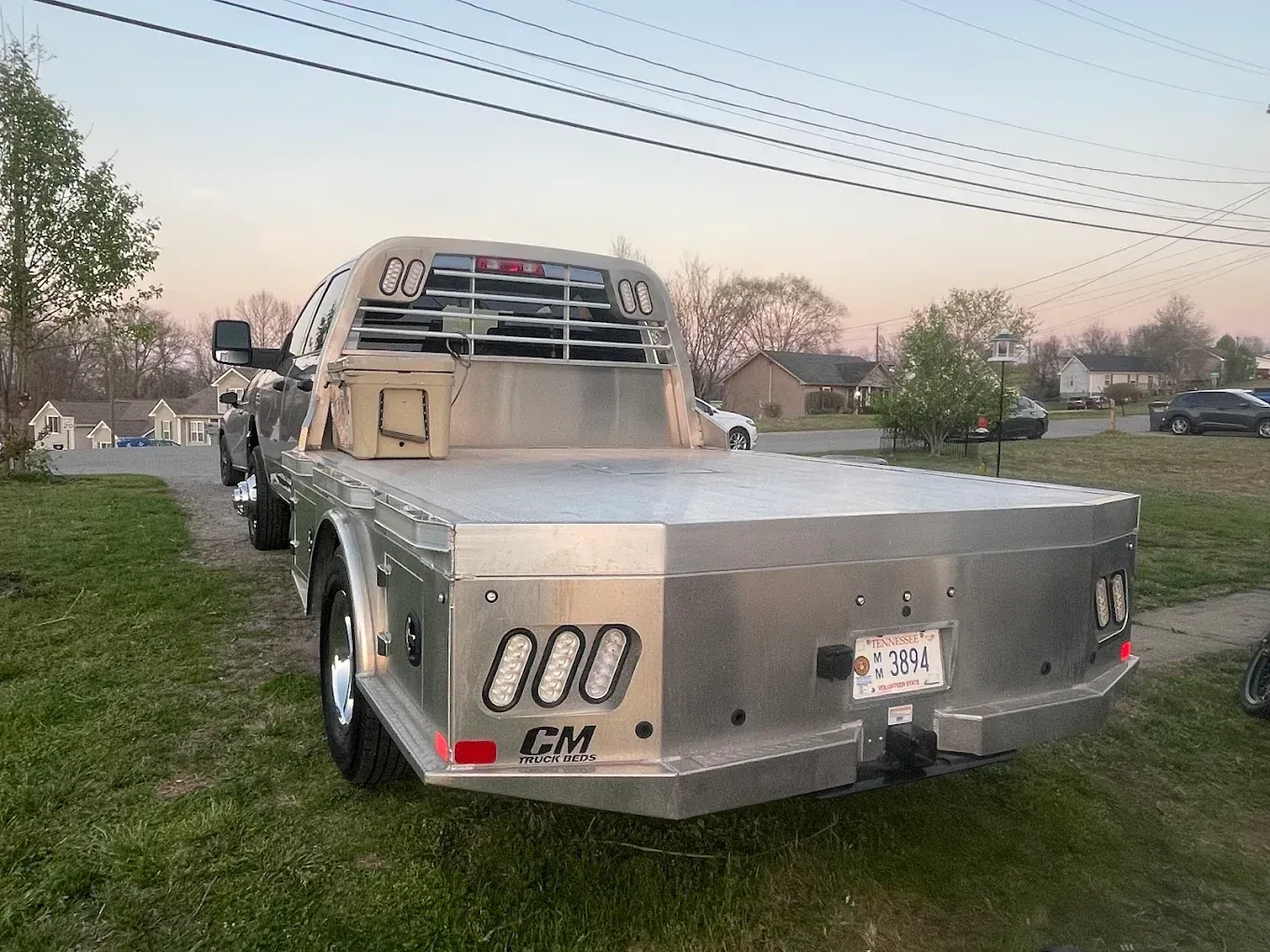Silver flatbed tow truck parked on grass at dusk, rear view with work lights and utility poles overhead