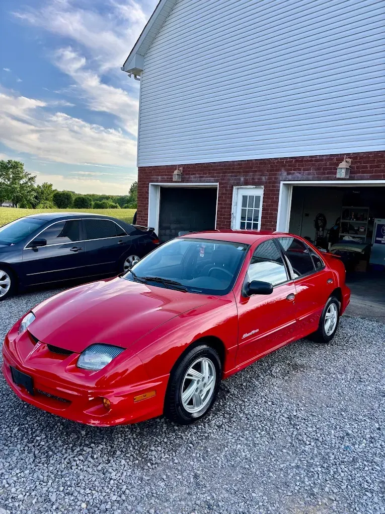 Red sports car parked on gravel beside a white barn with two garage bays, black car in background.
