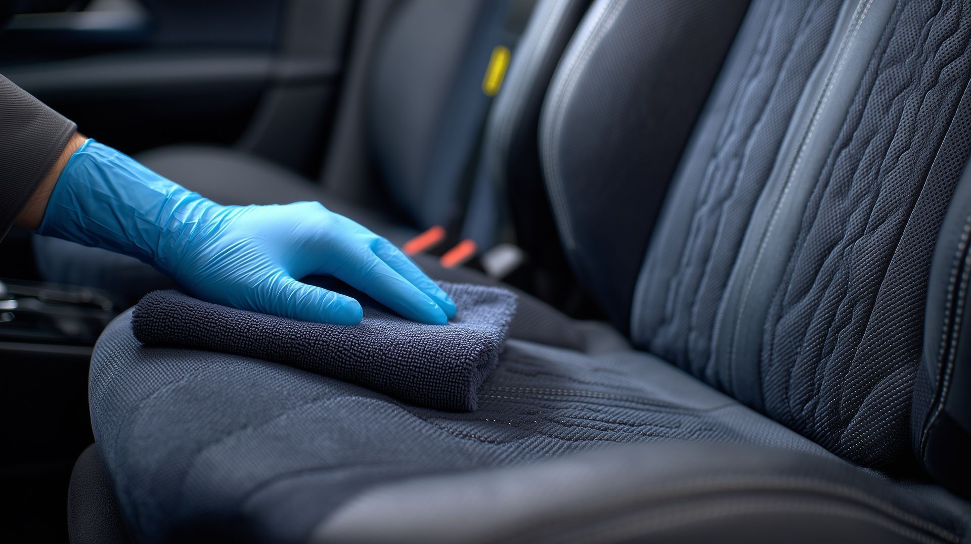 Blue-gloved hand cleaning a dark car seat with a cloth in the back seat area