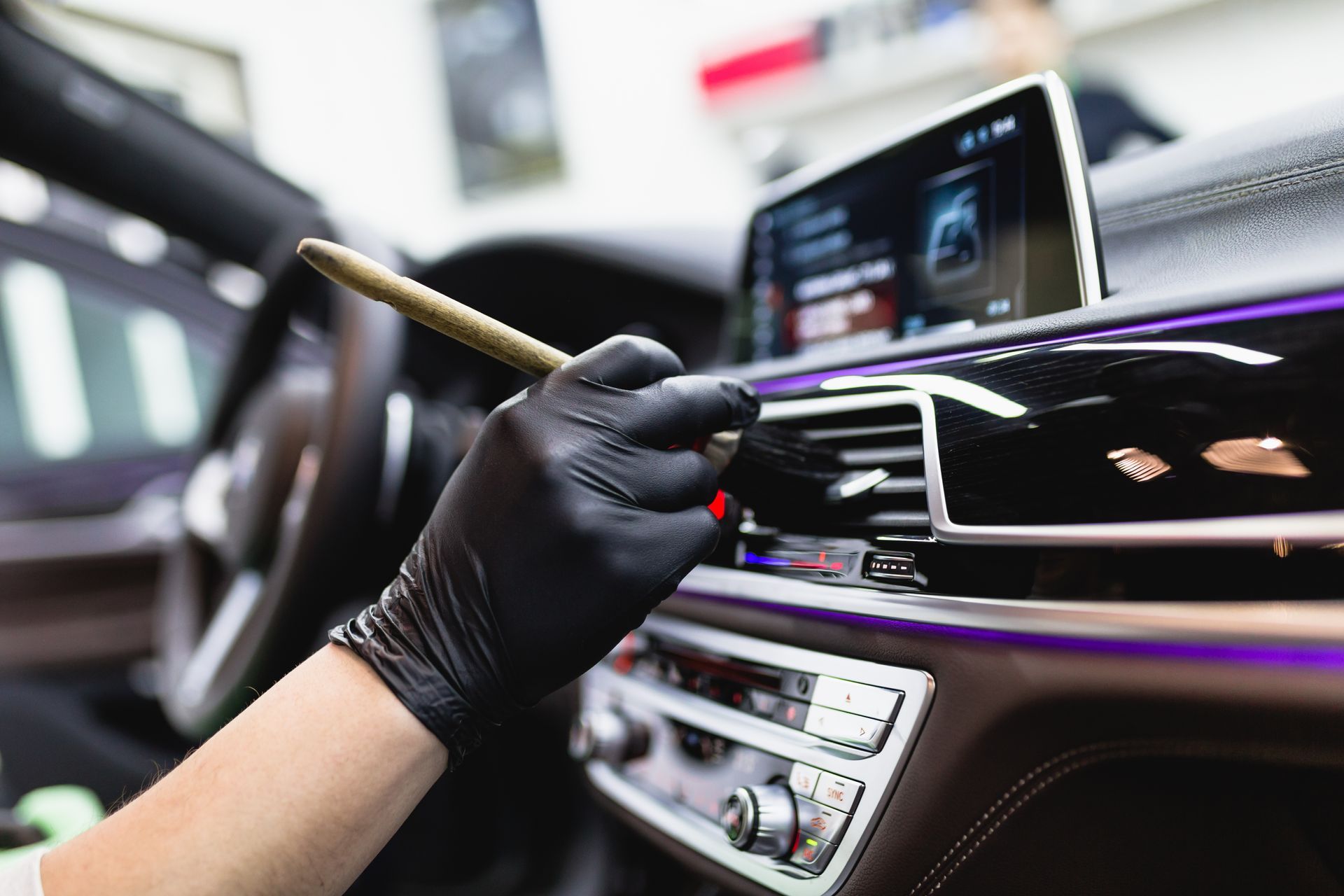 Gloved hand dusting a car dashboard with a brush, cleaning the interior near a touchscreen display