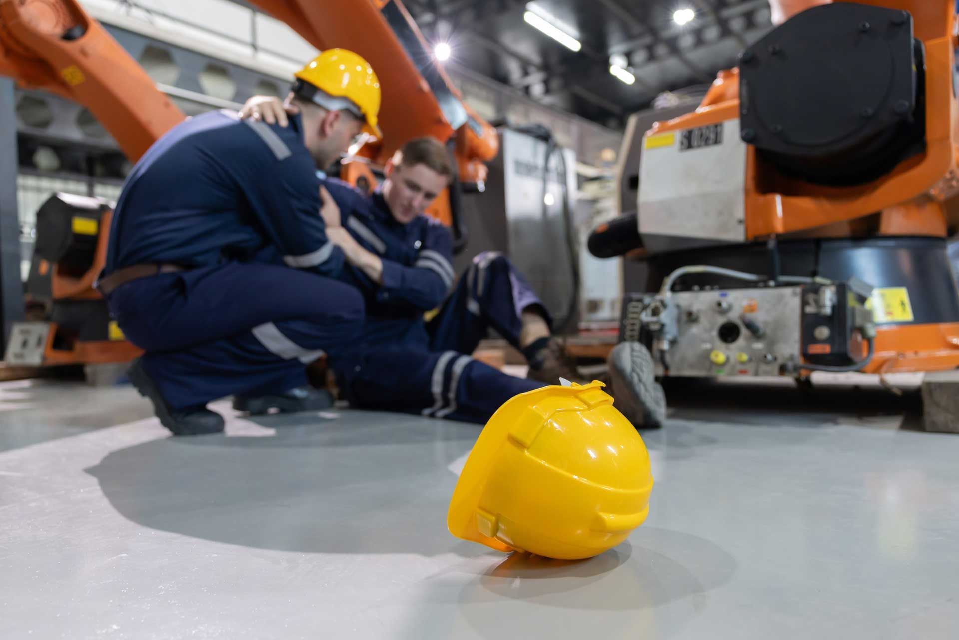 A close-up on a yellow hardhat with a worker helping an injured colleague on the floor of a factory.