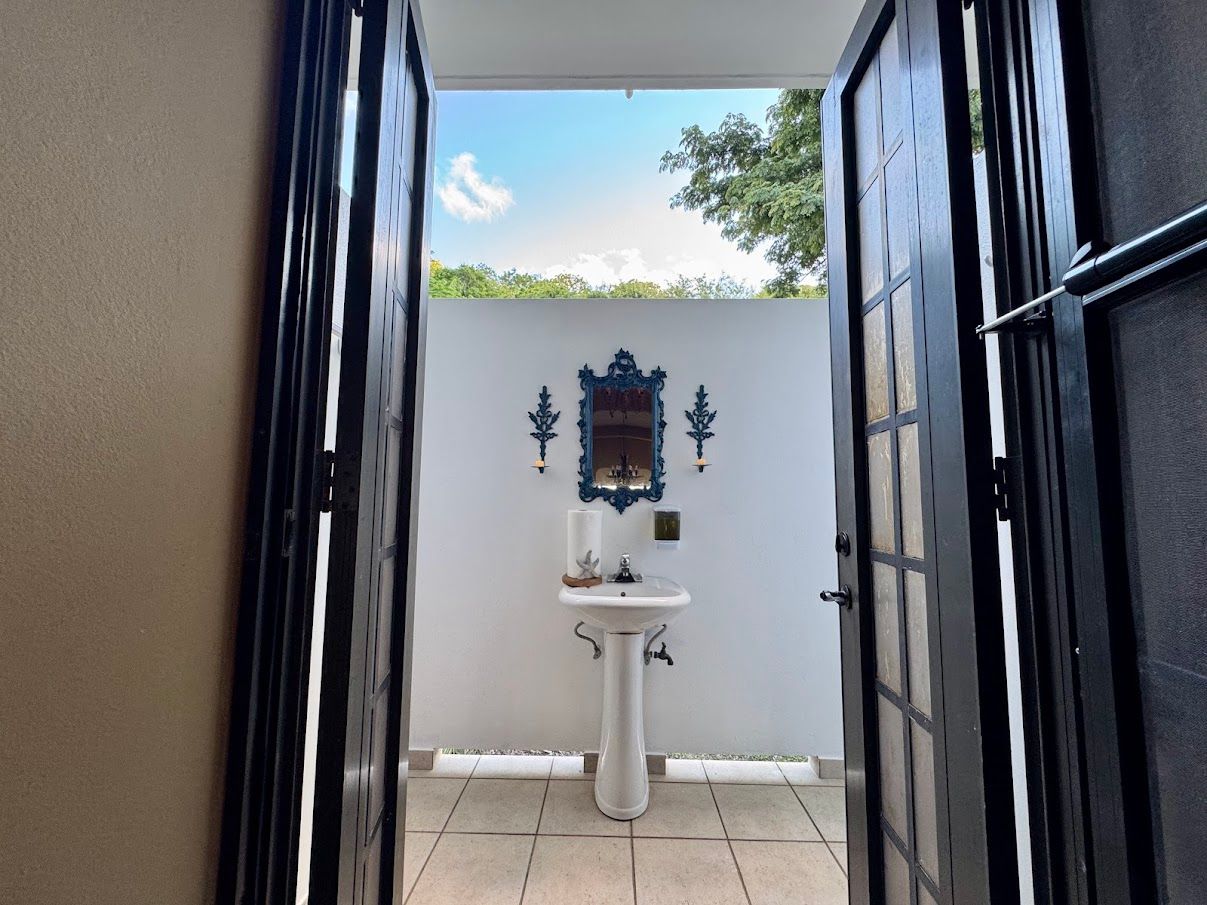 Open black doors frame a small outdoor sink area with a blue ornate mirror and blue sky view.