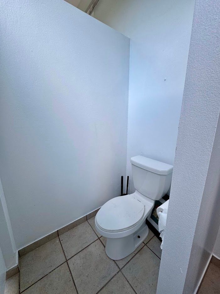 A white toilet in a small, white-walled bathroom with tan tile flooring.