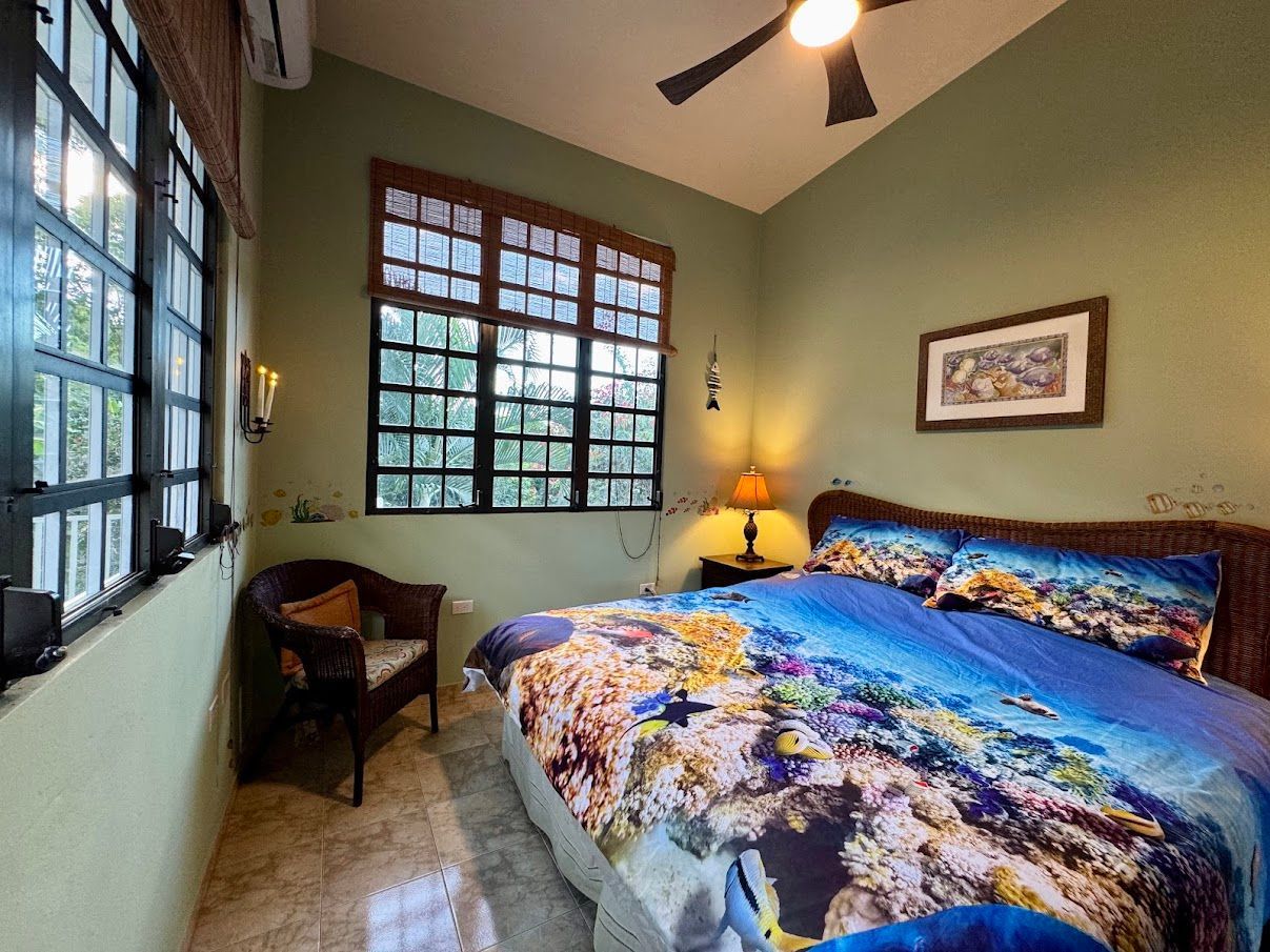 Bedroom with two windows, a bed with coral reef bedding, and a wicker chair.