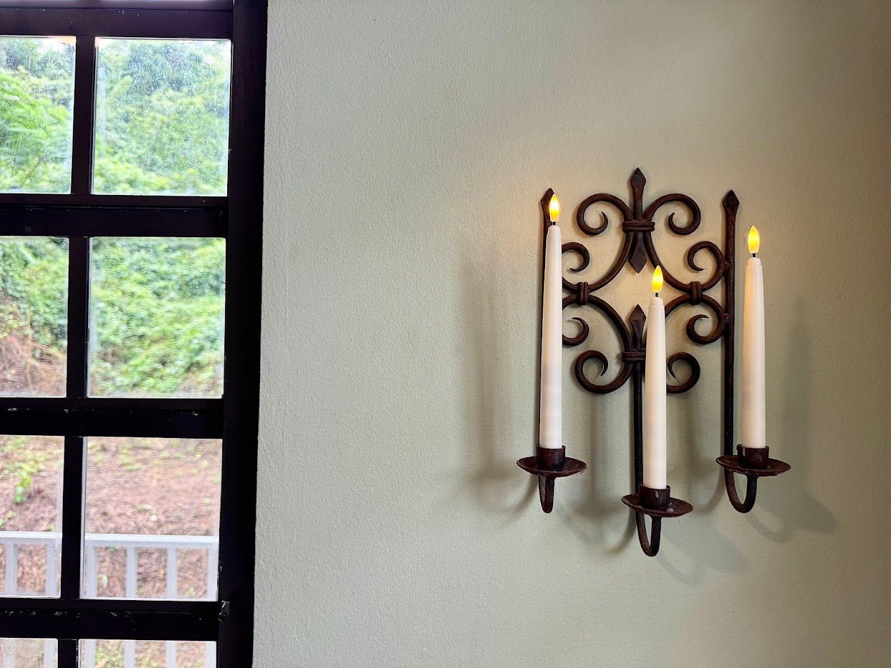 Black wrought iron candle holder with three lit candles on light green wall, window with greenery.