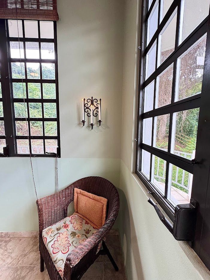 Wicker chair with floral cushion by windows. Candle sconce on light green wall. Exterior view of trees.