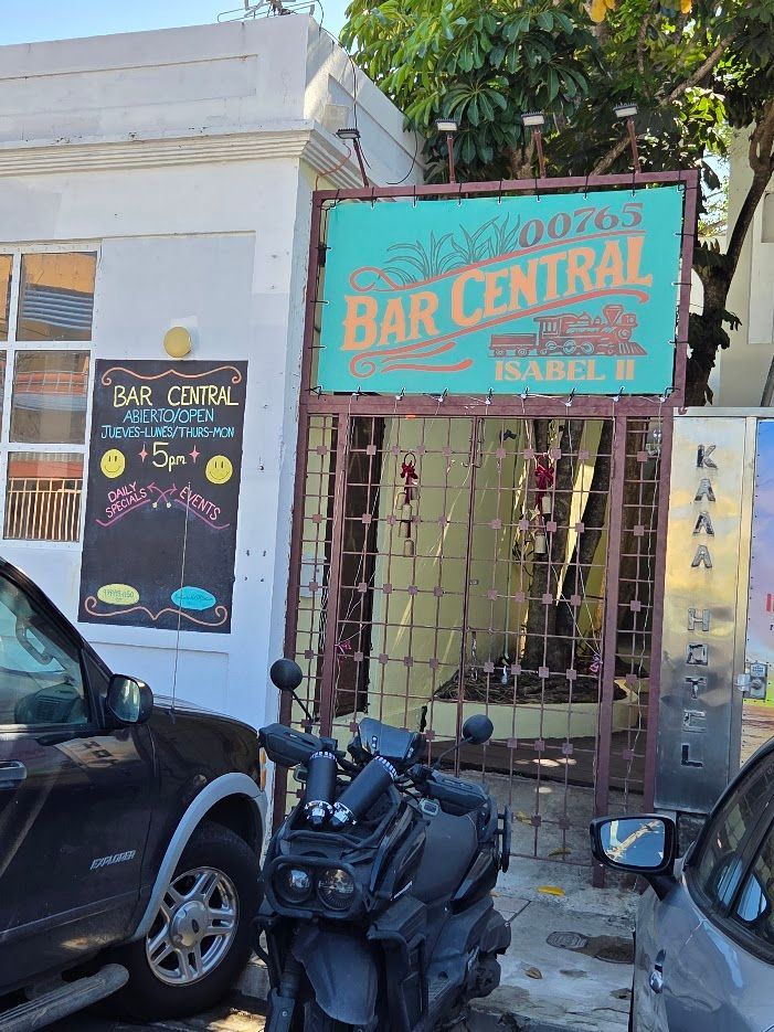 Interior of El Plaza restaurant, tables set for dining, pendant lights, wooden sign, plants, and natural light.