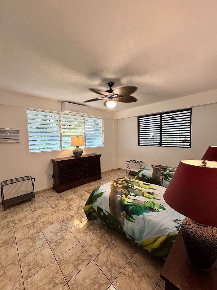 Bedroom with two beds, dresser, windows with blinds, ceiling fan, and luggage rack.